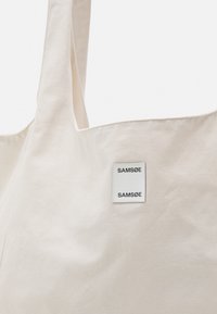 Beżowa torba zakupowa z płótna z szerokimi uchwytami oraz białą prostokątną etykietą z czarnym napisem "SAMSØE" z przodu.