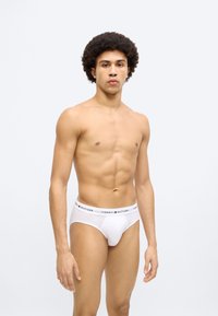 Λευκά σλιπ εσώρουχα με μαύρη και κόκκινη μέση "TOMMY HILFIGER". Ομαλή υφή, εφαρμοστό σχέδιο και κλασική κοπή. Το μοντέλο στέκεται σε ουδέτερη πόζα.