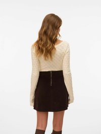 Maglione di tessuto crema con maniche lunghe, abbinato a una gonna in velluto a coste marrone scuro con zip centrale. I capelli cadono in onde morbide.