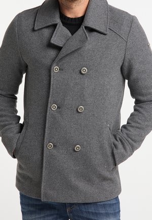 Homme portant un manteau en laine gris à double boutonnage avec six boutons métalliques et les mains dans les poches.