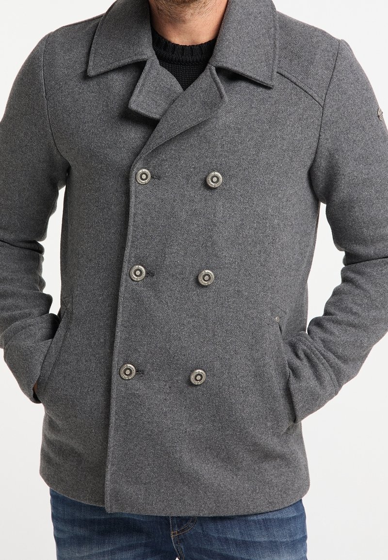 Homme portant un manteau en laine gris à double boutonnage avec six boutons métalliques et les mains dans les poches.