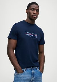 Ναυτικό μπλε βαμβακερό T-shirt με το λογότυπο "tommy" σε πολυχρωμο κείμενο, στρογγυλή λαιμόκοψη, κοντά μανίκια, και προβάλλεται με μπλε τζιν παντελόνι.