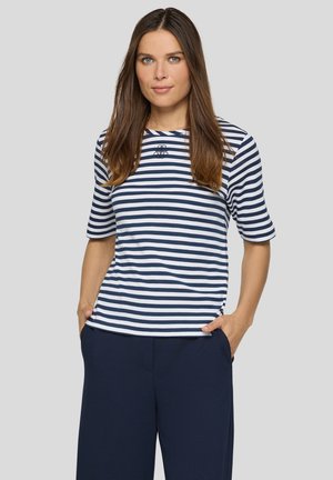 Frau mit langen braunen Haaren, die ein marineblau-weiß gestreiftes kurzärmeliges Oberteil und dunkelblaue Hose trägt, steht mit den Händen in den Taschen.