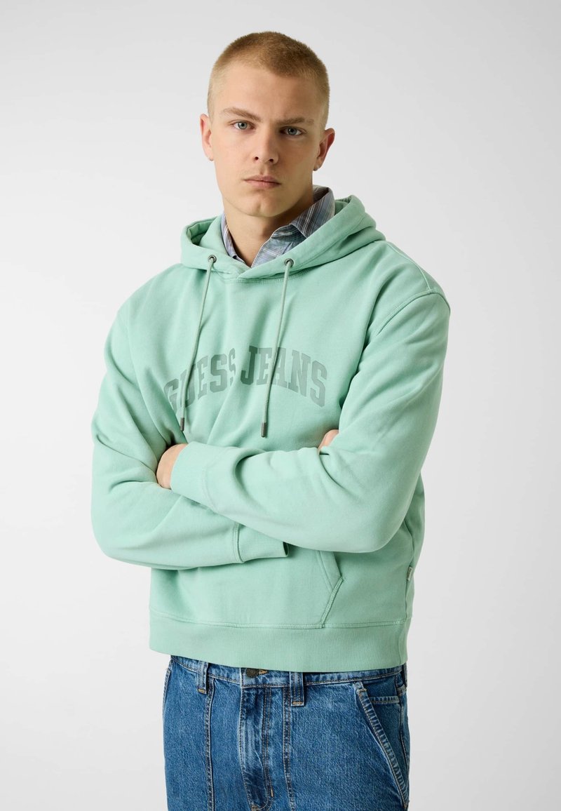 Jeune homme les bras croisés portant un sweat à capuche vert menthe Guess Jeans par-dessus une chemise à carreaux et un jean bleu, debout devant un fond uni.