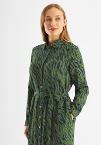 Rochie din cămașă cu mânecă lungă, de culoare verde, cu un model ondulat în nuanțe mai închise, având guler, închidere cu nasturi și o curea legată în talie pentru a contura silueta.
