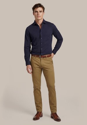 Man draagt een marineblauw overhemd met lange mouwen en knopen, beige chino, bruine riem en bruine nette schoenen, staand tegen een effen beige achtergrond.