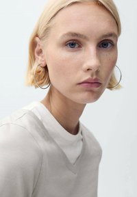 Prim-plan al unui model cu păr scurt blond, purtând un top gri deschis cu decolteu în V. Cercei mari din argint accentuează trăsăturile ei pe un fundal deschis.
