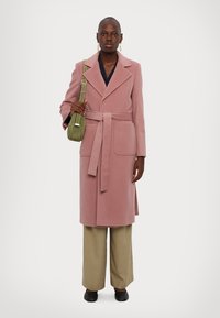 Lång rosa kappa med snedskurna slag, midjebälte och två framfickor, i kombination med vida beiga byxor och en liten grön axelväska.