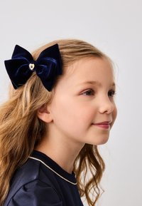 Fascetta di velluto navy con un centro a forma di cuore dorato, indossata su capelli mossi. In primo piano su uno sfondo neutro, che mette in risalto la texture dell'accessorio.