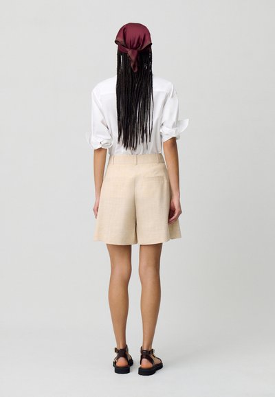 Blusa bianca con maniche arrotolate, pantaloni corti beige e sandali marroni. La sciarpa è bordeaux. I pantaloni corti sono tagliati, di lunghezza media fino alla coscia e con tasche.
