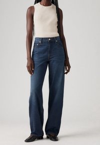 Person med en ärmlös beige ribbad topp, hög midja med vida mörkblå jeans och svarta lågklackade skor på en enfärgad bakgrund.