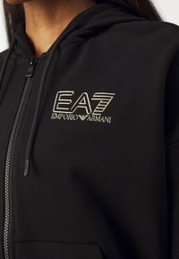 Črna zip-up kapuca iz mehkega blaga. Ima zlat logo EA7 na prsnem delu, kapuco z vrvicami in kengurujskimi žepi.