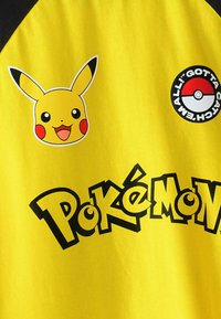 Keltainen T-paita, jossa on Pikachu-grafiikka hymyilevällä naamalla ja mustilla yksityiskohdilla, sekä pyöreä Pokéball-logo ja "Pokémon"-teksti.