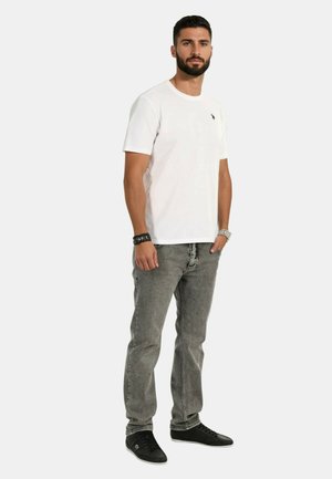 Ein weißes T-Shirt mit kurzen Ärmeln, das ein kleines Logo aufweist. Kombiniert mit grauen Jeans und schwarzen Sneakers entsteht ein lässiger, moderner Look.