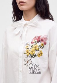Femme portant une chemise blanche boutonnée avec un nœud au col et une poche à motif floral avec des fleurs jaunes, blanches et roses et le texte "MG LISA".