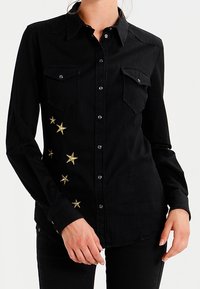Camicia nera con bottoni, due tasche sul petto e ricamo a stella dorato sul lato sinistro, realizzata in tessuto morbido con colletto classico.