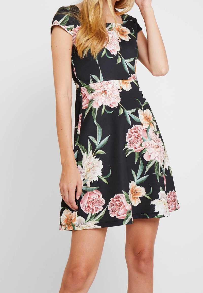 Femme portant une robe noire à manches courtes avec des motifs floraux roses et blancs, debout avec une main près du visage et l'autre détendue.