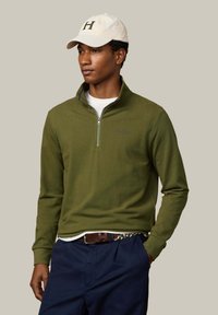 Sudadera con cremallera en cuello color verde oliva con logo, combinada con una camiseta blanca. También incluye pantalones azul oscuro y una gorra de béisbol de color claro.