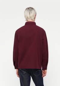 Burgunderfarvet langærmet skjorte med krave, knappede manchetter og en afslappet pasform. Stoffet ser glat ud. Set fra ryggen, parret med mørk denim.