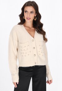 Cardigan beige realizzato in morbido tessuto, con scollatura a V, polsini a costine e due tasche frontali decorate con pietre decorative e bottoni.