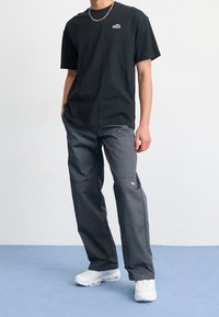 Camiseta de algodón negra con un pequeño logo, combinada con pantalones cargo grises de corte holgado que presentan bolsillos y un parche con la marca, complementados con zapatillas blancas.