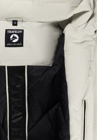 Beige en zwarte gewatteerde jas met het label "TRAVELIN' waar we lopen" en maat M-tag binnenin de kraag, zijrits zak zichtbaar op de mouw.