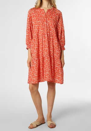 Mujer con vestido naranja holgado con estampado abstracto blanco, mangas tres cuartos y sandalias planas beige, de pie sobre un fondo liso.