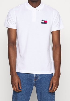 Polo bianca in cotone con colletto classico. Presenta una toppa colorata con il logo "TOMMY JEANS" sul lato sinistro del petto. Vestibilità casual.