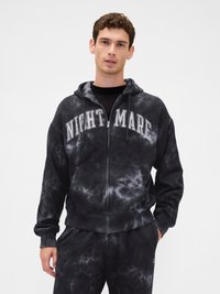 Jovem vestindo um hoodie com estampa tie-dye em preto e cinza, com o texto "NIGHT MARE", uma camisa preta por baixo e calças também com estampa tie-dye, em fundo branco.