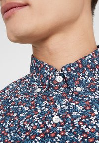 Camicia con motivo floreale che presenta uno sfondo blu navy con fiori bianchi, rossi e blu. Il colletto è abbottonato con bottoni bianchi.