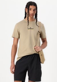 Beige bavlněné tričko s černým logem "Timberland", spárované s černými šortkami a béžovou kabelkou na rameno. Volný střih a minimalistický design.