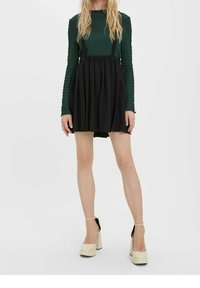 Zwarte geplisseerde rok met verstelbare bretels, gecombineerd met een getextureerde groene lange mouw top. Model draagt crèmekleurige plateauhakken.