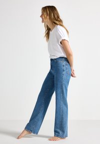 Jeans in denim blu a vita alta e gamba larga con un lavaggio sbiadito, abbinati a una maglietta bianca semplice. A piedi nudi, in posizione laterale.