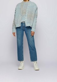 Lichtblauwe gebreide cardigan met een gestructureerd patroon, grijze trui eronder, gecombineerd met een rechte spijkerbroek en witte hoge sneakers.