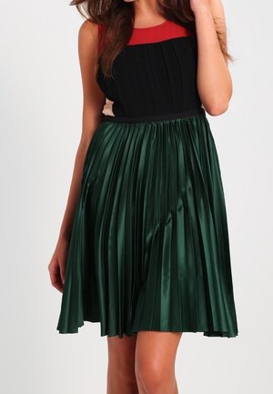 Plisowana sukienka z czarnym gorsetem i czerwonym akcentem, z zieloną satynową spódnicą. Spódnica ma gładką teksturę i sięga tuż nad kolano.