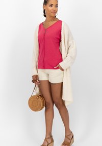 Roze top met korte mouwen en knoopdetails, crèmekleurige gebreide cardigan, witte shorts en een ronde rieten tas. Model draagt tan gekleurde plateau-sandalen.