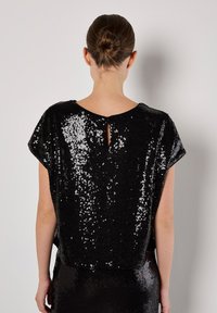 Zwarte pailletten top met een losse pasvorm, voorzien van een ronde halslijn en een keyhole detail op de rug. Glanzend oppervlak dat licht reflecteert.