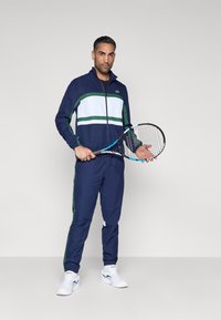 Chaqueta azul marino con cremallera y franjas verdes y blancas, pantalones a juego azul marino y zapatillas deportivas blancas. El modelo sostiene una raqueta de tenis.