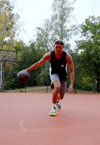 Giocatore di basket che dribbla una palla su un campo rosso, indossando una maglietta senza maniche nera, pantaloni grigi e scarpe nere e bianche con dettagli verdi.
