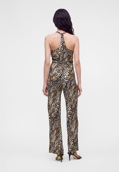 Femme debout, de dos, portant une combinaison sans manches à col halter avec un motif abstrait zèbre marron, noir et blanc, et des talons hauts noirs.