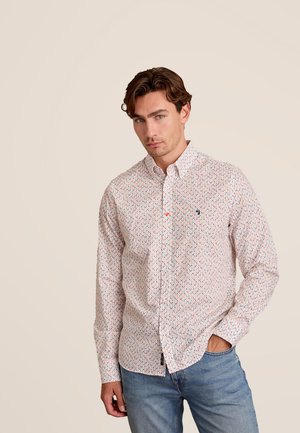 Witte overhemdd met kleurrijk geometrisch patroon, kraag en enkele zak. Gepaard met lichtblauwe spijkerbroek tegen een neutrale achtergrond.