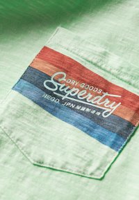 Primer plano de tela verde claro con un bolsillo cosido que presenta franjas horizontales en rojo, naranja, azul marino y azul, y texto del logotipo "Superdry" en blanco.