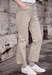 Personne portant un pantalon en velours côtelé beige, les mains dans les poches, des chaussettes grises et des baskets blanches, debout sur une rue pavée.