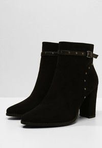 Botas de tobillo de ante negro con puntera puntiaguda, superficie texturizada y suela con tacón. Presentan una correa de cuero con tachuelas y acentos metálicos.