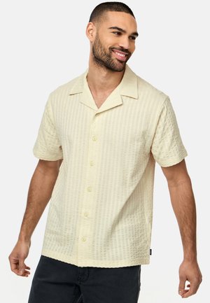 Homme souriant aux cheveux courts et à la barbe, portant une chemise beige texturée à manches courtes avec boutons et un pantalon noir, debout devant un fond blanc.