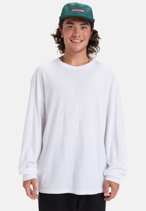 White long-sleeve shirt with a relaxed fit and crew neckline, featuring subtle embroidered detailing at the center. Paired with a green cap.