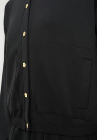 Μαύρο φούτερ φτιαγμένο από μαλακό υλικό με χρυσά κουμπιά, δύο μπροστινές τσέπες και ελαστική μέση για εφαρμοστή εμφάνιση.