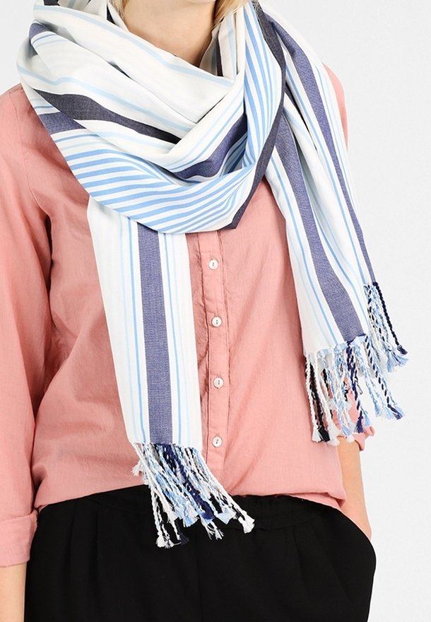 Femme portant une grande écharpe blanche avec des rayures bleu et bleu marine et des franges, par-dessus une chemise rose boutonnée et un pantalon noir.
