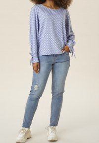 Blusa azzurro chiaro con motivo, maniche lunghe e dettagli legati ai polsini, abbinata a jeans blu strappati e sneaker bianche.