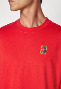 Punainen puuvillapaita, jossa on pyöreä kaulus, ribbauskaulus ja brodeerattu monivärinen Nike-logo-merkki vasemmassa rinnassa.